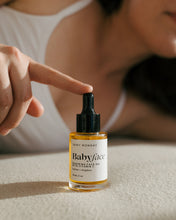 Load image into Gallery viewer, A baby face soothing face oil bottle with a black dropper cap and white label, placed on a beige surface. A person in a white tank top points at the bottle with their index finger.
